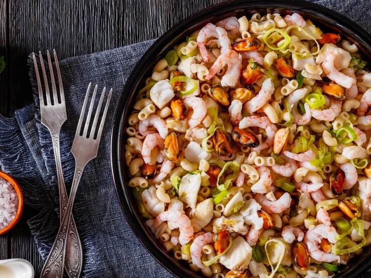 Makkaroni-Meeresfruechtesalat Eine schwarze Schüssel mit dem Salat auf einem dunklen Holztisch. Daneben noch einige Gewürze und eine graue Tischdecke mit zwei Gabeln.