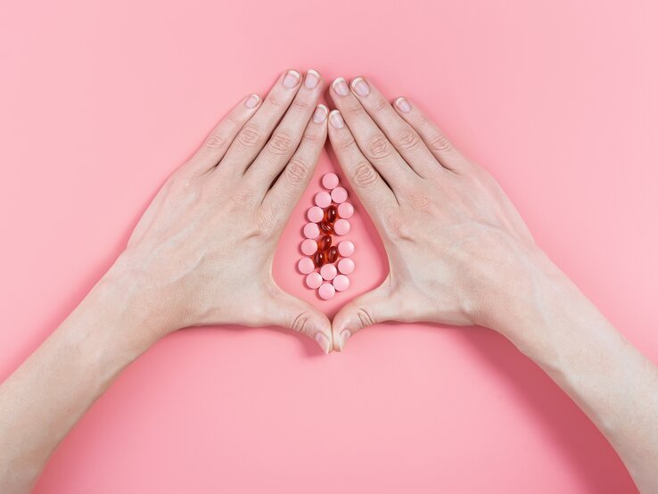 Zwei Frauenhände umschließen vor einem rosa Hintergrund eine Ansammlung von Pillen, so dass das Gebilde aussieht wie eine Vagina.