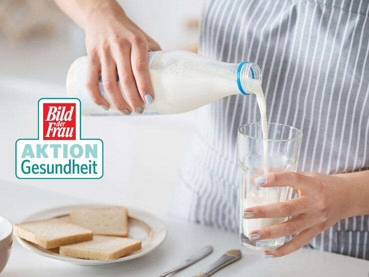 Eine Frau im gestreiften Oberteil gießt frische Milch aus einer Glasflasche in ein klares Glas. Die Szene spielt in einer hellen Küche, was auf eine alltägliche Haushaltstätigkeit hindeutet. Im Hintergrund ist ein Teller mit Scheiben Weißbrot und eine Schale zu sehen, was auf ein bevorstehendes Frühstück schließen lässt. Auf dem Bild ist ebenfalls das "Bild der Frau AKTION Gesundheit" Logo zu erkennen. Die gepflegten Hände der Frau und die sorgfältige Ausführung der Handlung vermitteln ein Gefühl von Fürsorge und Gesundheitsbewusstsein.