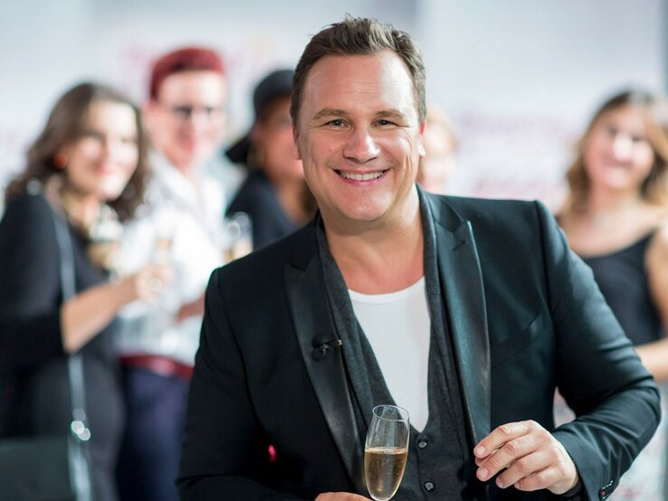 Ein lächelnder Guido Kretschmer in einem schwarzen Blazer und weißem T-Shirt hält ein Glas Sekt und blickt in die Kamera. Er steht im Vordergrund, während im Hintergrund unscharf weitere Personen zu sehen sind, die ebenfalls Getränke halten und sich in einer offensichtlich geselligen Atmosphäre befinden.