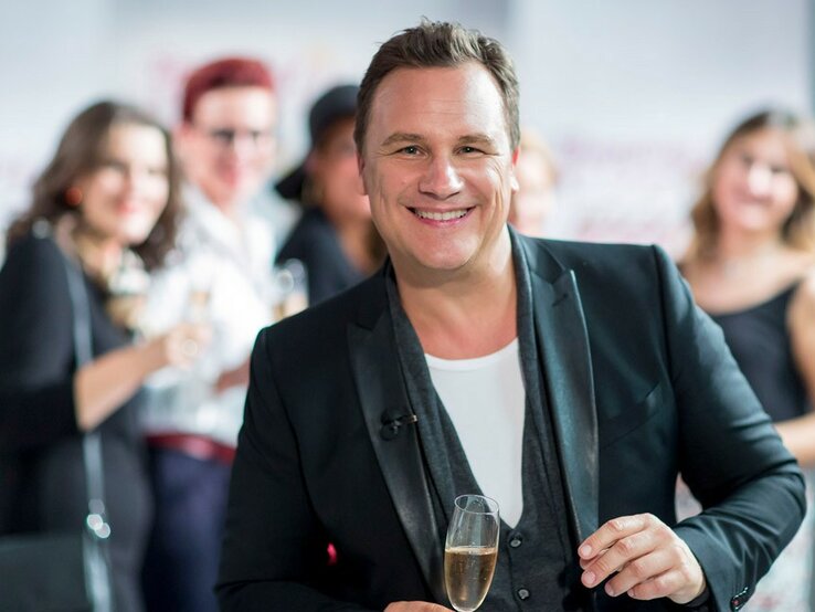 Guido Maria Kretschmer Shopping Queen Ein lächelnder Guido Kretschmer in einem schwarzen Blazer und weißem T-Shirt hält ein Glas Sekt und blickt in die Kamera. Er steht im Vordergrund, während im Hintergrund unscharf weitere Personen zu sehen sind, die ebenfalls Getränke halten und sich in einer offensichtlich geselligen Atmosphäre befinden.