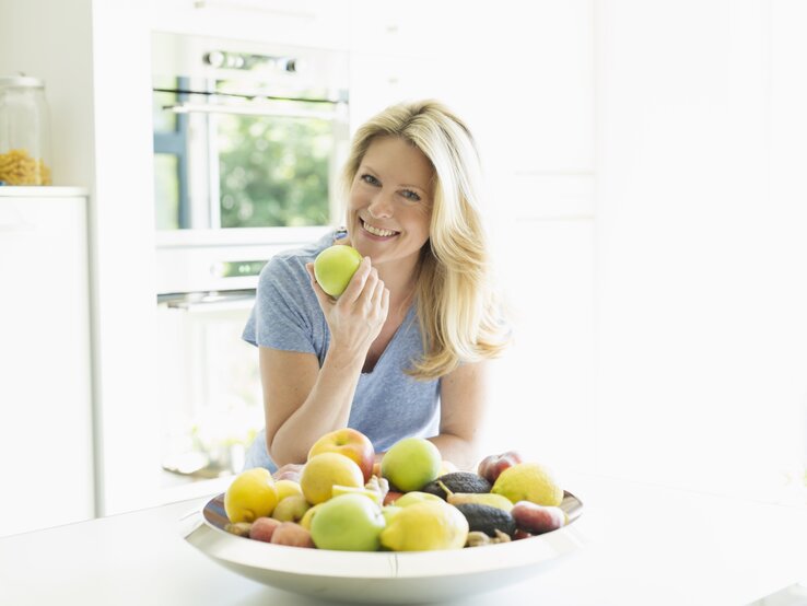 Eine lächelnde blonde Frau hält einen grünen Apfel in ihrer Hand und steht vor einem Tisch mit einer Schale voller frischer, bunter Früchte. Sie trägt ein blaues T-Shirt und befindet sich in einer hellen, sonnendurchfluteten Küche, was ein gesundes und glückliches Lebensgefühl vermittelt.
