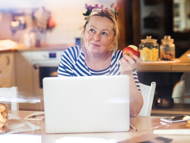 Reife Frau sitzt an einem Laptop und hat einen Apfel in der Hand. Sie sieht sehr zuversichtlich aus. 