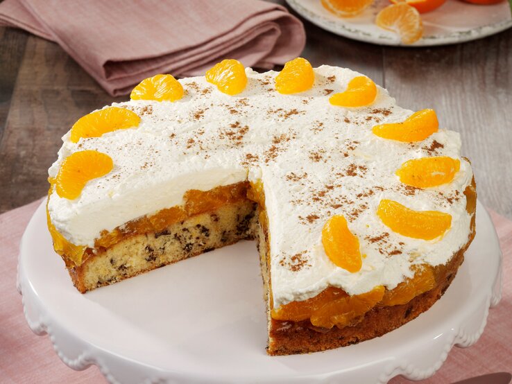 Eine Mandarinen-Sahnetorte mit Schokoraspeln und Schmand auf einem Kuchentablett. Die Torte ist bereits angeschnitten und zwei Stücke wurden heraus genommen.