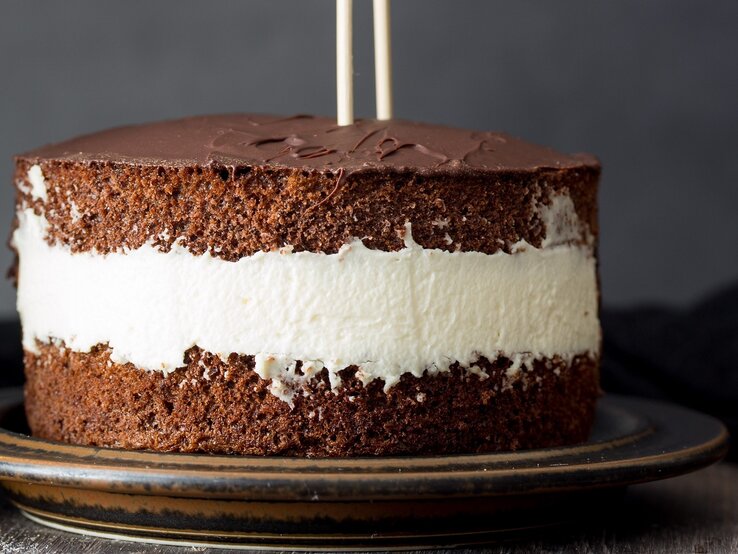 Die Torte auf einem dunkelbraunen Teller und dunklem Hintergrund mit zwei Mikadostäbchen in der Torte. Daneben ein Löffel und eine schwarze Tischdecke.