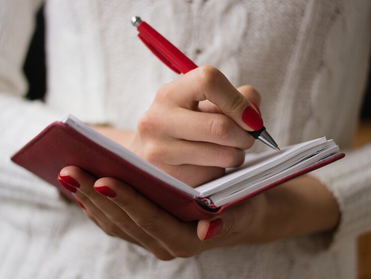 Frau mit roten Fingernägeln schreibt mit einem Kugelschreiber handschriftlich in ein Notizbuch. 