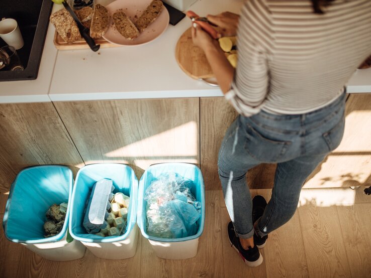 Eine Frau steht in der Küche und schält Karotten und Kartoffeln. Die Reste wandern in die neben ihr stehenden Mülltonnen.
