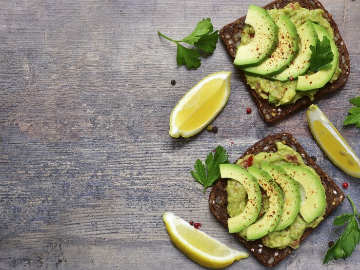 Zwei dunkle Brotscheiben mit Avocados belegt direkt von oben auf einem dunklen Tisch fotografiert.