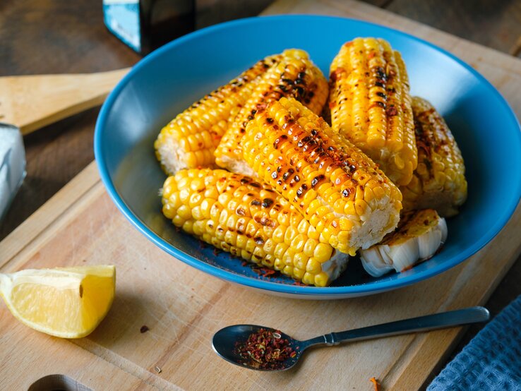 Das Bild zeigt eine Nahaufnahme von gegrillten Maiskolben, die appetitlich in einer tiefen blauen Schüssel serviert werden. Die Maiskörner haben eine leuchtend gelbe Farbe mit Grillspuren, die ihnen ein rustikales Aussehen verleihen. Neben der Schüssel liegt ein Stück Zitrone, dessen frische Farbe einen schönen Kontrast zum Gelb des Maises bildet. Auf dem Schneidebrett vor der Schüssel ist ein Löffel mit einer Gewürzmischung zu sehen, was darauf hindeutet, dass der Mais gewürzt wurde. Im Hintergrund sind Küchenutensilien und eine klare Flasche, möglicherweise Öl oder Essig, angedeutet, was die häusliche und gemütliche Atmosphäre unterstreicht.