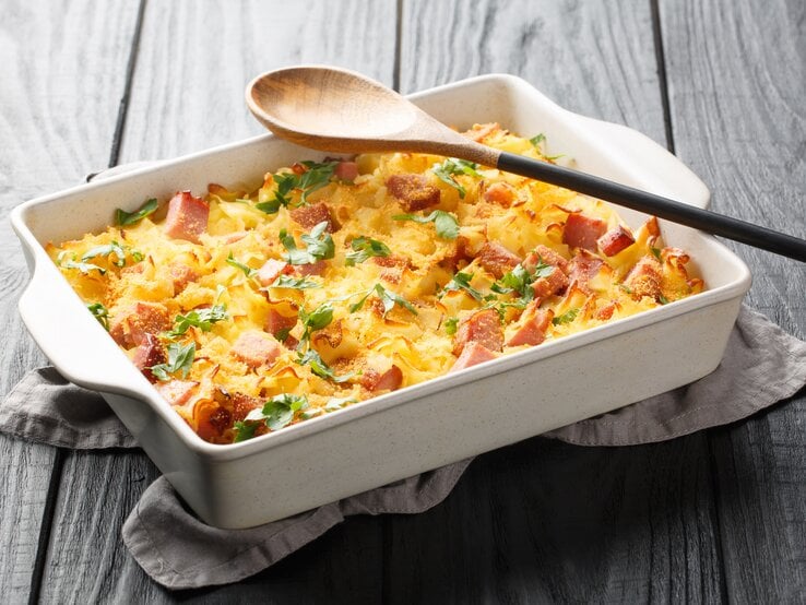 Schinken-Fleckerl-Auflauf in weißer Auflaufform auf dunklem Holz. Auf der Form liegt ein Holzkochlöffel.
