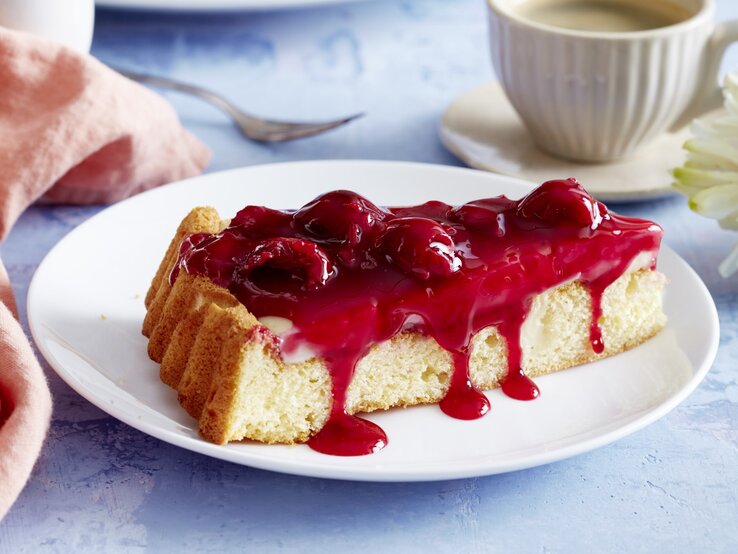Unser Pudding-Kirschkuchen schmeckt wirklich jedem! Pudding-Kirschkuchen auf einem weißen Teller, blauer Untergrund und Geschirr und Küchentuch im Hintergrund.