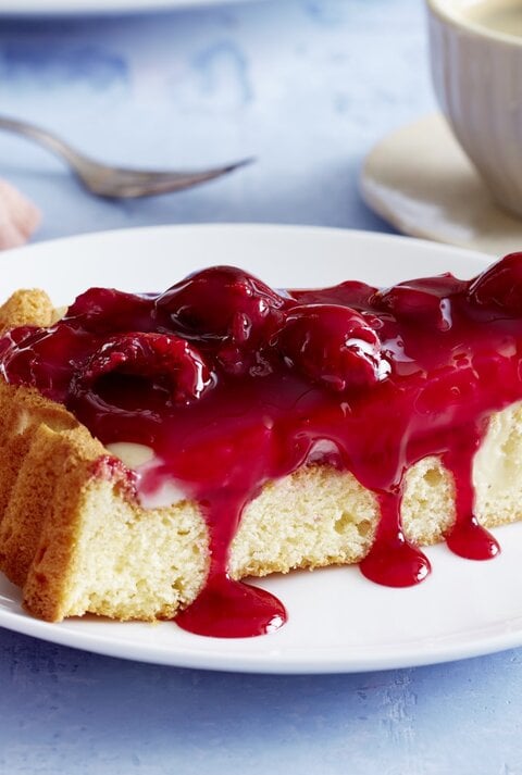 Pudding-Kirschkuchen auf einem weißen Teller, blauer Untergrund und Geschirr und Küchentuch im Hintergrund. 