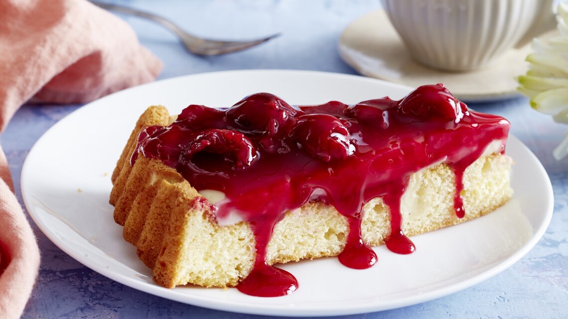 Pudding-Kirschkuchen auf einem weißen Teller, blauer Untergrund und Geschirr und Küchentuch im Hintergrund. 