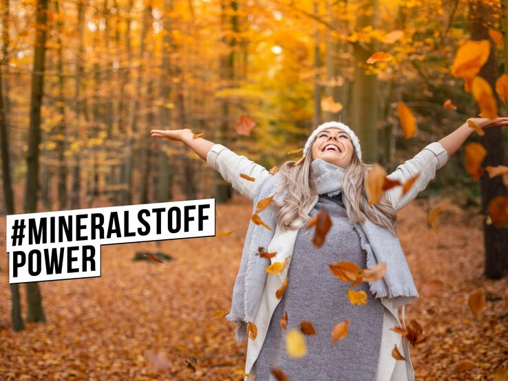 Eine glücklich lächelnde Frau mit grauem Hut und Schal streckt die Arme aus und genießt den Herbst im Wald. Sie ist umgeben von fallenden Blättern und einem Hintergrund aus leuchtend orangefarbenen Bäumen. Die Frau trägt eine graue Strickjacke und scheint den Moment der Ruhe in der Natur zu genießen. Über ihr ist das Hashtag #MINERALSTOFFPOWER in schwarzer Schrift auf einem weißen Balken abgebildet.