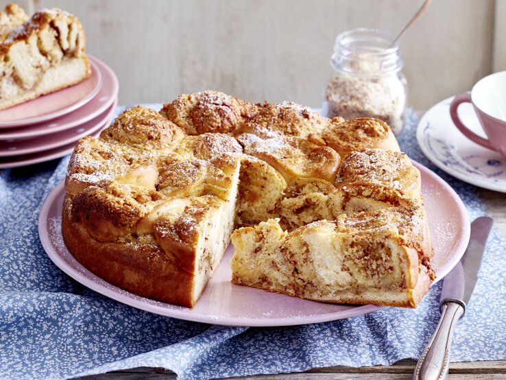 Ein Dänischer Butterkuchen steht auf einem Tisch. Daneben liegen mehrere Teller, ein Messer sowie eine Tasse mit einem Unterteller.