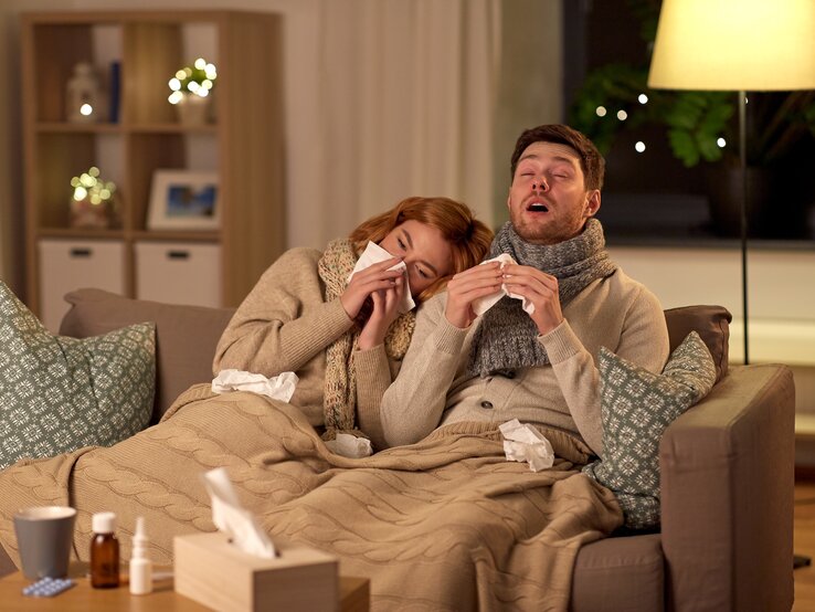Ein junges Paar sitzt abends erkältet auf dem Sofa, er niest gerade, während sie sich an ihn angelehnt die Nase putzt. Beide sind in Schals, Strickjacken und Decken eingewickelt. 