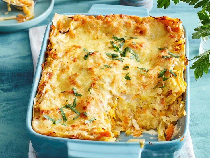 Blaue Ofenform mit angeschnittener Gemüse-Lasagne, Geschirr und Kräuter im blauen Hintergrund.