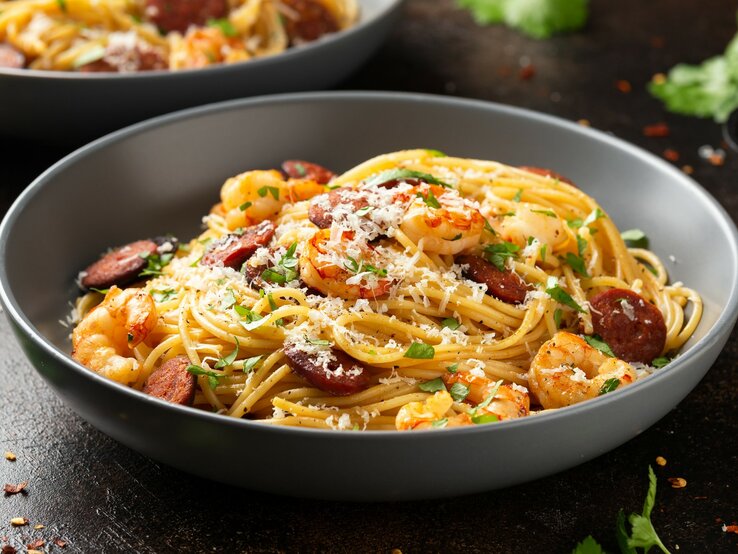 Ein grauer Teller mit der Garnelen- und Chorizopasta auf einem dunklen Holztisch mit vielen Kräutern und Käse bestreut.