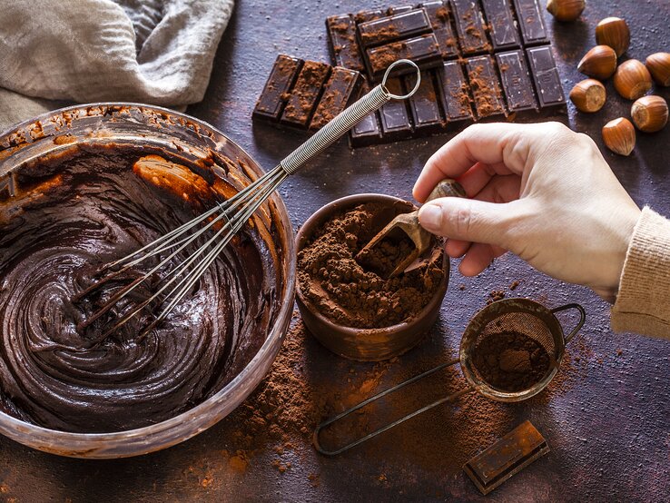 Dunkler Untergrund bedeckt mit allerlei Zutaten für einen Schokoladenkuchen, dessen Teig gerade in einer Schüssel angerührt wird. 