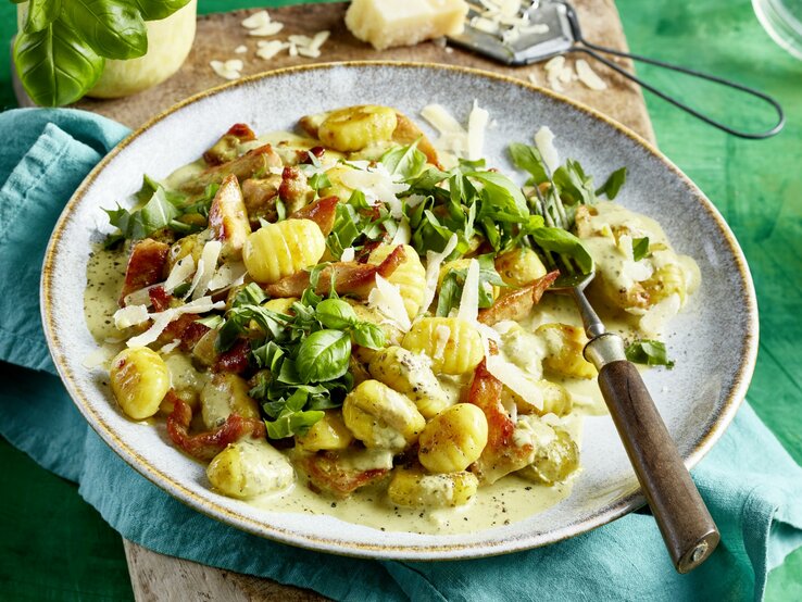 Pesto-Geschnetzeltes mit Gnocci, frischem Rucola und Basilikum und geriebenem Parmesan, serviert auf einem tiefen Keramikteller auf grünem Untergrund. 