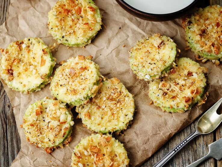 Zucchini-Parmesan-Chips auf einem Backpapier. Daneben Besteck und ein Ausschnitt einer Schale.