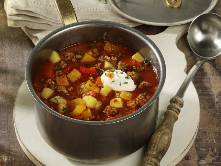 Bauerneintopf mit Hackfleisch, Paprika ud Kartoffeln, garniert mit Schmand und frischem Schnittlauch, serviert in einem großen Topf auf einem rustikalen Holztisch. 