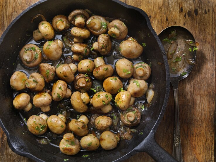 Das Bild zeigt eine gusseiserne Pfanne mit gebratenen Champignons, die auf einem rustikalen Holztisch steht. Die Pilze sehen saftig aus und sind mit Kräutern, wahrscheinlich Petersilie, bestreut