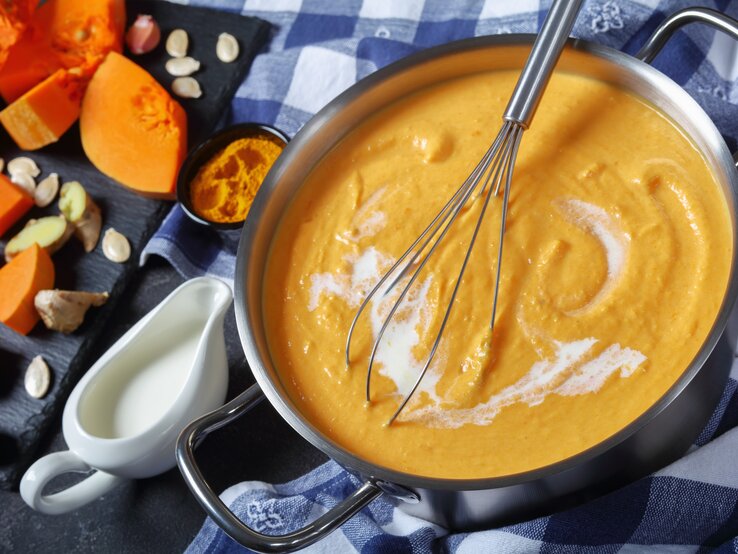 Ein Topf mit der Kürbissuppe auf einem Tisch mit blau-weiß-karierter Tischdecke. 