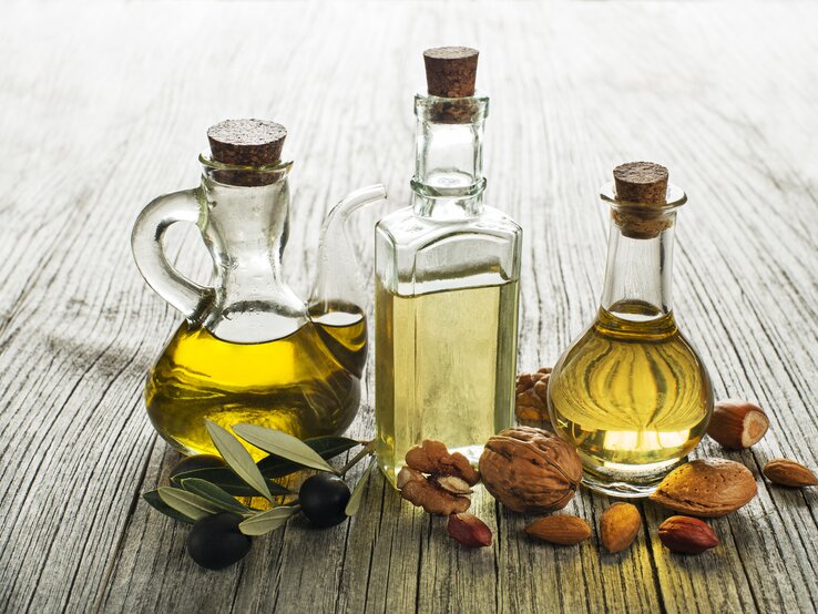 Drei Glasflaschen mit Olivenöl und Essig umgeben von Oliven und Nüssen auf rustikalem Holztisch.