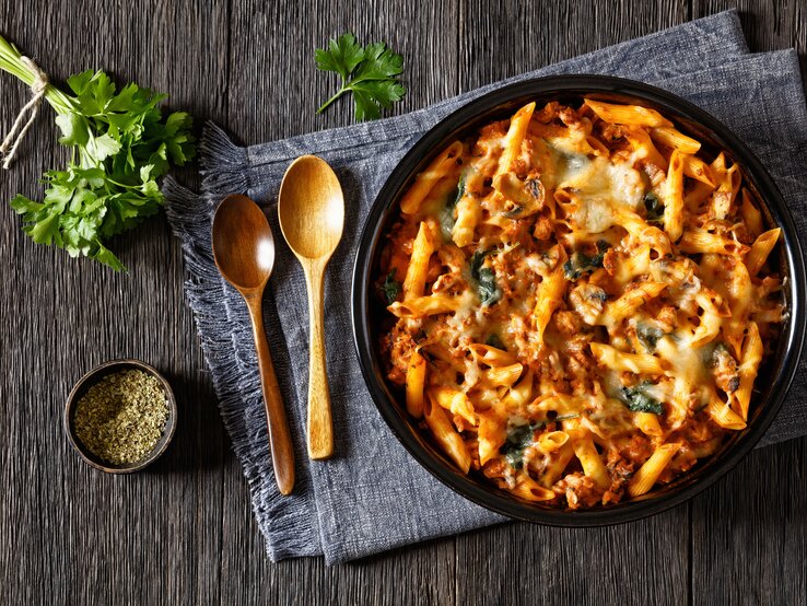 Gebackene Hähnchen-Nudeln mit Spinat und Pilzen in Tomatensoße auf schwarzem Teller. Dieser steht auf einem mit einem Geschirrtuch bedeckten Holztisch. Daneben liegen Holzlöffel, Petersilie und eine schale schwarzer, gemahlener Pfeffer.