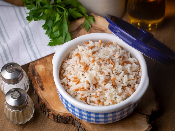 Draufsicht: In einer Porzellanschüssel ist gekochter Reis. Auf dem Gefäß ist der Deckel angelehnt. Das Gefäß steht auf einem Holzbrett. Daneben stehen ein Salz- und ein Pfefferstreuer.