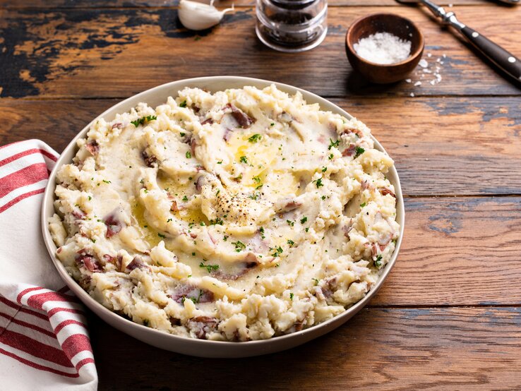 Frontalsicht: eine große Schale mit einem Knoblauch-Kartoffelpüree aus roten Kartoffeln steht auf einem Untergrund aus Holz. Daneben liegen ein Handtuch, Besteck, eine Knoblauchzehe und ein Schälchen mit Salz.