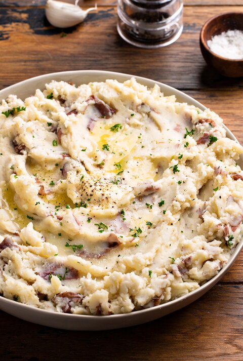 Frontalsicht: eine große Schale mit einem Knoblauch-Kartoffelpüree aus roten Kartoffeln steht auf einem Untergrund aus Holz. Daneben liegen ein Handtuch, Besteck, eine Knoblauchzehe und ein Schälchen mit Salz.