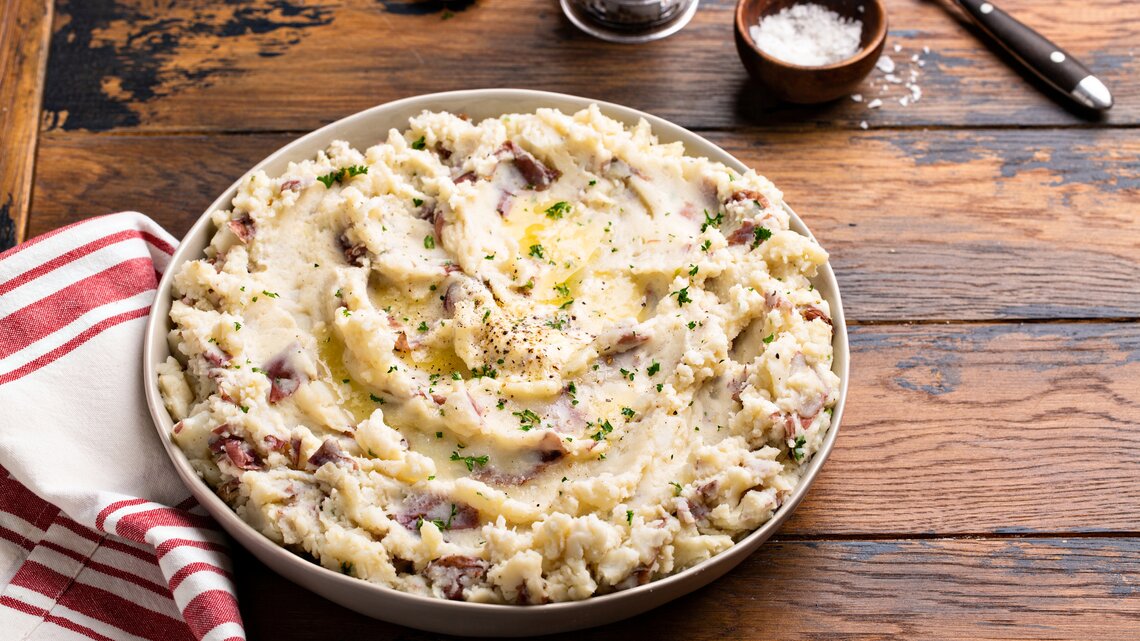 Frontalsicht: eine große Schale mit einem Knoblauch-Kartoffelpüree aus roten Kartoffeln steht auf einem Untergrund aus Holz. Daneben liegen ein Handtuch, Besteck, eine Knoblauchzehe und ein Schälchen mit Salz.