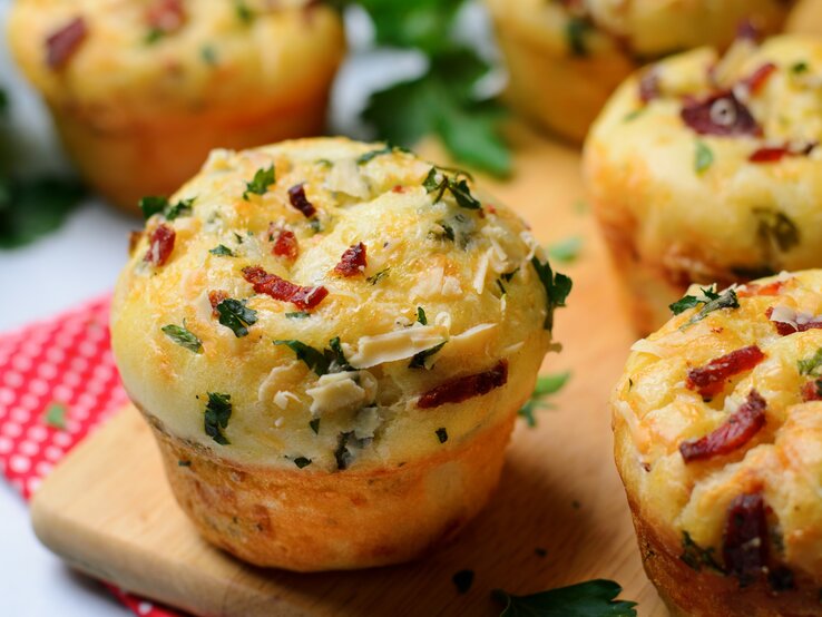 Einige Pizza-Muffins auf einem Holzschneidebrett und darunter eine rot-weiße Decke. Heller Hintergrund und einige Kräuter sind auch noch zu sehen.