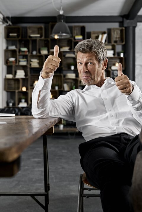 Mann in einem modernen Büro. Er sitzt lässig auf einem Stuhl, hat seine Füße auf den Tisch gelegt und zeigt mit beiden Daumen nach oben. Er trägt ein weißes Hemd und eine dunkle Hose. Seine Körperhaltung und der Gesichtsausdruck vermitteln Selbstbewusstsein und Gelassenheit.