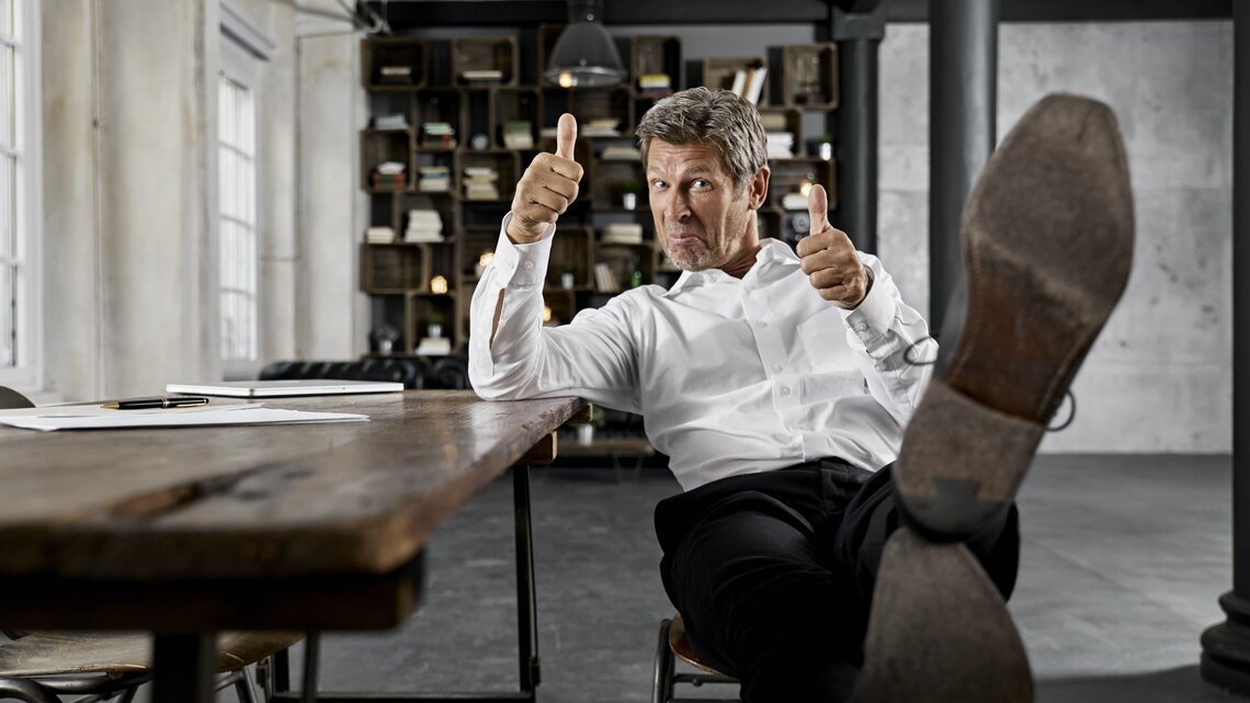 Mann in einem modernen Büro. Er sitzt lässig auf einem Stuhl, hat seine Füße auf den Tisch gelegt und zeigt mit beiden Daumen nach oben. Er trägt ein weißes Hemd und eine dunkle Hose. Seine Körperhaltung und der Gesichtsausdruck vermitteln Selbstbewusstsein und Gelassenheit.