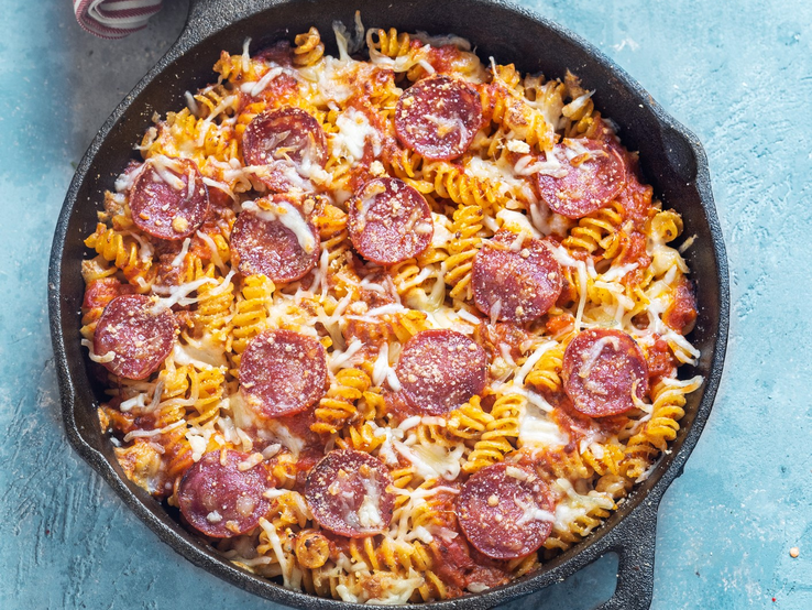 Spirelli-Salami-Pfanne mit Tomaten in Gusspfanne auf blauem Untergrund. Oben im Bild rot-weiß-gestreiftes Tuch. Aufnahme aus der Vogelperspektive.