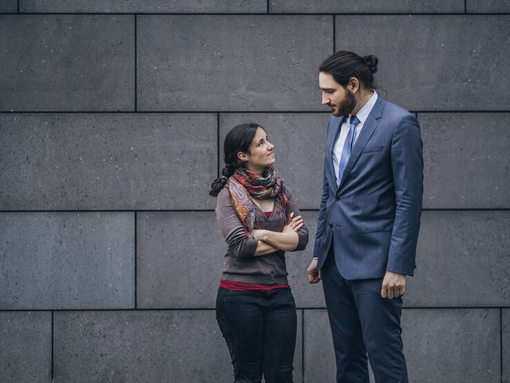 Mann und Frau sehen sich leicht lächelnd, aber etwas skeptisch an, sie ist sehr leger gekleidet, er trägt einen Anzug. Die beiden stehen vor einer Betonwand.