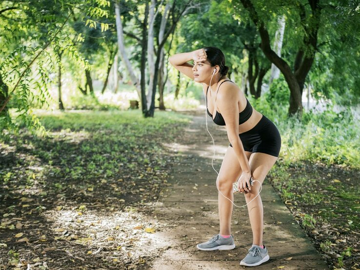 Eine Frau in Sportbekleidung macht eine Pause beim Joggen in einem grünen Park. Sie hält sich die Hand an die Stirn, hat Kopfhörer in den Ohren und sieht erschöpft aus.