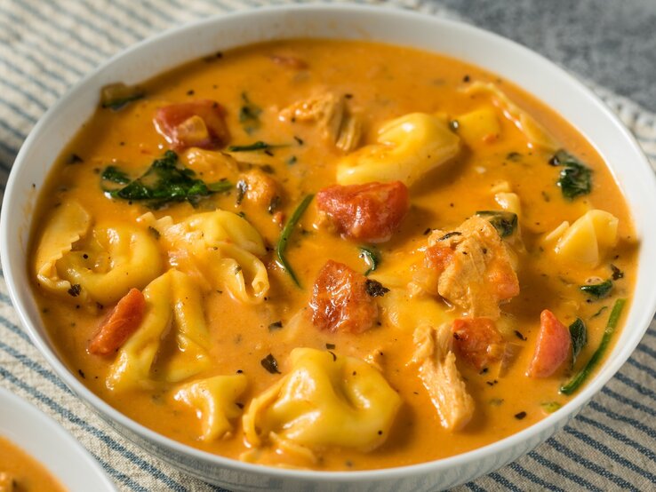 Ein weißer Teller mit der Tortellini-Suppe auf einem grauen Untergrund mit einem blau-weiß-gestreiften Tuch