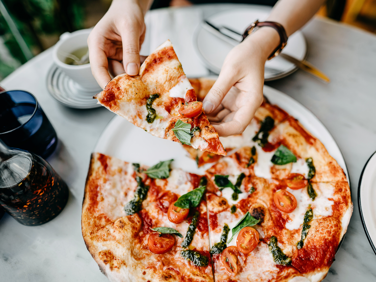 Das Bild zeigt eine Person, die ein Stück Pizza Margherita anhebt. Die Pizza ist belegt mit geschmolzenem Mozzarella, frischen Basilikumblättern und halbierten Kirschtomaten auf einer dünnen, leicht gebräunten Kruste. Die Szene spielt sich in einem Restaurant ab, erkennbar an den weiteren Tellern und Gläsern auf dem Tisch sowie dem Olivenöl und dem Gewürzstreuer im Hintergrund, was eine gemütliche Essatmosphäre suggeriert. Die frischen Zutaten und die Art, wie die Pizza serviert wird, lassen darauf schließen, dass es sich um eine traditionell italienische Zubereitungsweise handelt.