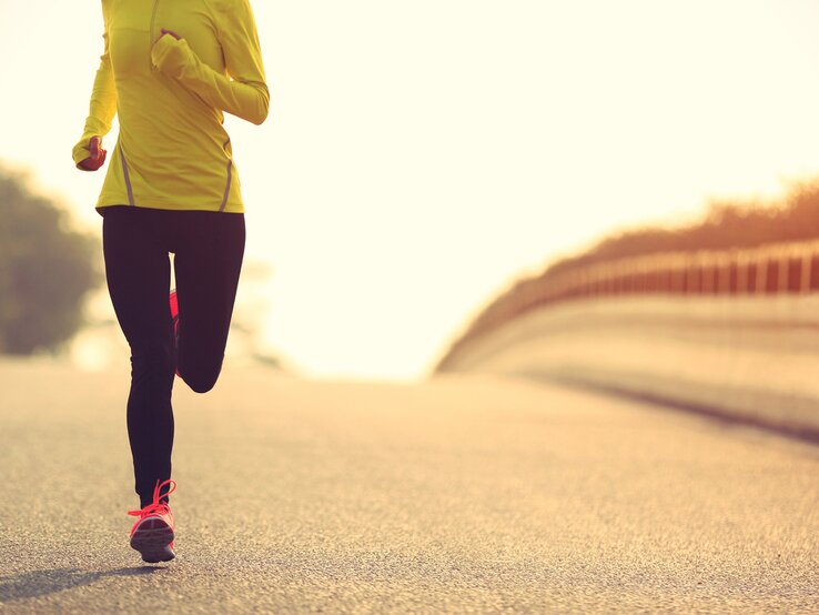  Ein Läufer in auffälliger Sportbekleidung joggt auf einer Straße, umgeben von sanftem Sonnenlicht und einer ruhigen Landschaft, was auf Training bei Tagesanbruch oder Sonnenuntergang hindeutet.