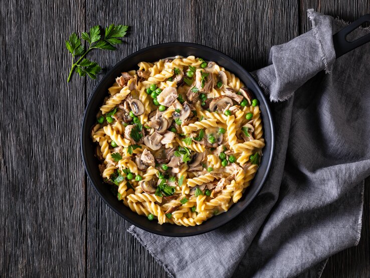 Thunfisch-Nudelpfanne mit Pilzen und Erbsen in Pfanne auf dunklem Untergrund. 