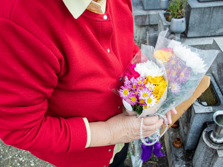 Eine ältere Frau steht mit Blumen in der Hand auf einem Friedhof an einem Grab
