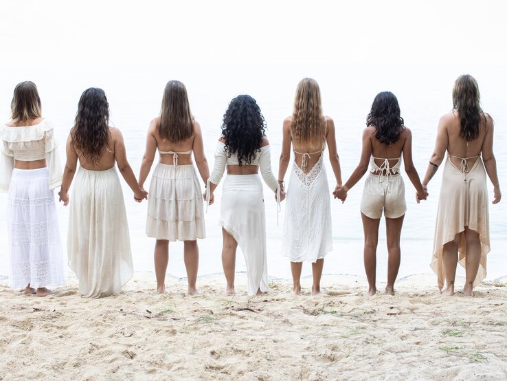 Sechs Frauen, die sich an den Händen halten und zum Meer hin ausgerichtet sind. Sie stehen auf Sand, was auf einen Strand hindeutet. Alle Frauen tragen weiße oder hellfarbene Sommerkleidung. Sie stehen mit dem Rücken zur Kamera. | © iStock.com/Jasmina007