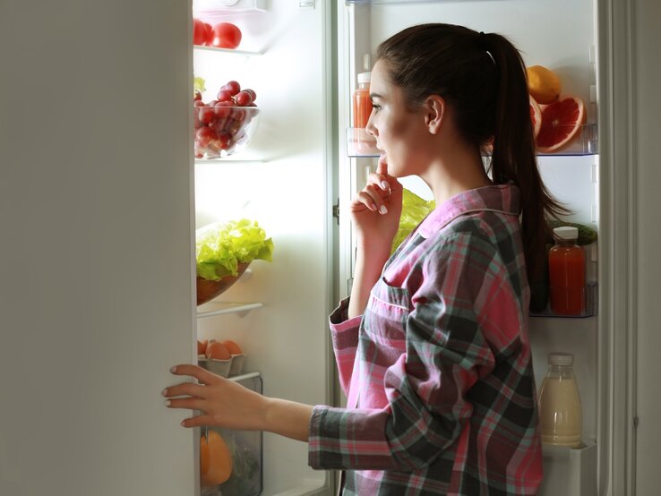 Eine junge, dunkelhaarige Frau, die nachts Hunger spürt, steht an der offenen Kühlschranktür und überlegt, was sie essen kann.
