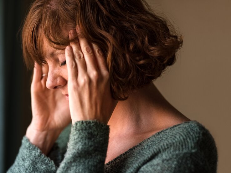  Das Bild zeigt eine Frau mittleren Alters, die sich gestresst oder besorgt zu fühlen scheint. Sie hat ihre Augen geschlossen und ihre Hände an die Schläfen gelegt, was oft eine Geste ist, die Menschen verwenden, wenn sie Kopfschmerzen haben, sich konzentrieren oder versuchen, sich zu beruhigen. Ihr Gesichtsausdruck deutet auf Unbehagen oder Schmerz hin. Sie trägt einen gemütlichen, grünen Pullover, und der Hintergrund ist unscharf, was darauf hinweist, dass der Fokus auf ihrem emotionalen Zustand liegt.