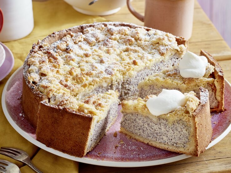 Auf einem Teller ist ein Streusel-Mohn-Käsekuchen serviert. Er ist mit Puderzucker bestreut und wird mit einem Klecks Sahne serviert.