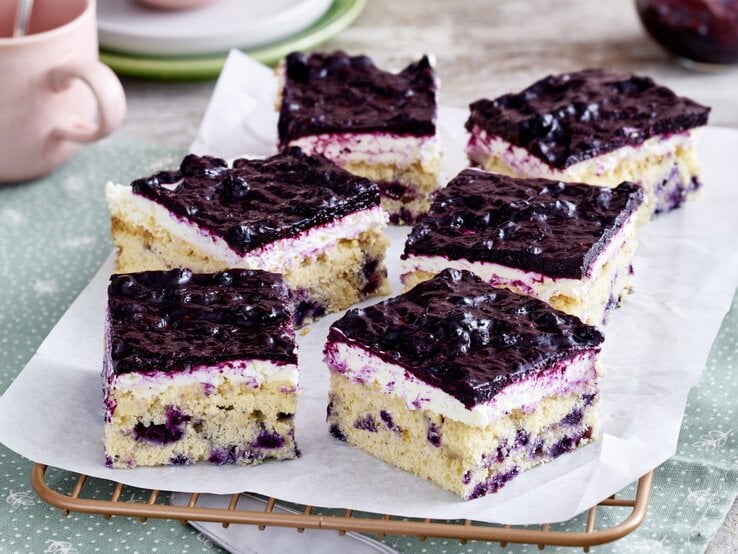 Sechs viereckige Stücke Heidelbeer-Quarkkuchen auf einem Backpapier auf Kuchengitter mit hellgrünem Untergrund. Im Hintergrund Geschirr. 